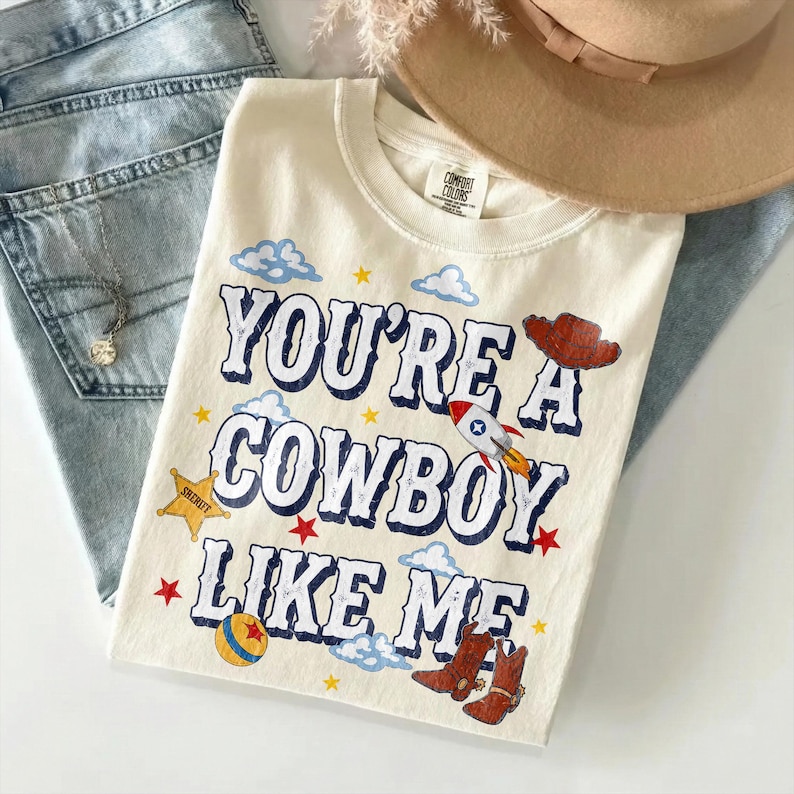 Puede incluir: Una camiseta de color crema con el texto "YOU'RE A COWBOY LIKE ME" en letras azules. El dise&ntilde;o incluye ilustraciones de un sombrero de vaquero marr&oacute;n, un cohete, una estrella de sheriff amarilla, una pelota colorida y botas marrones.