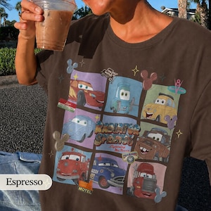 May include: A dark brown t-shirt featuring a grid graphic of characters from the movie Cars, including Lightning McQueen, Mater, and others, with the text Radiator Springs, Lightning McQueen, and various small icons like Mickey Mouse ears.