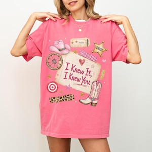 May include: A bright pink t-shirt featuring a graphic print of a cowboy hat, a wagon wheel, a sheriff badge, a cowboy boot, and a lined notepad with the text "I Knew It, I Knew You" surrounded by stars, a heart, and a leopard print strip.