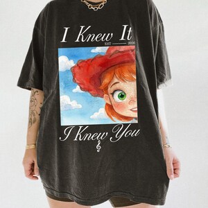 K&ouml;nnte beinhalten: Ein anthrazitfarbenes Kurzarm-T-Shirt mit einem Grafikdruck einer Zeichentrickfigur mit roten Haaren und einem roten Cowboyhut vor einem blauen Himmel mit Wolken. Der Text lautet I Knew It EST 2026 oben und I Knew You mit einem Violinschl&uuml;ssel unten.