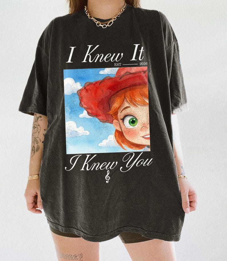 K&ouml;nnte beinhalten: Ein anthrazitfarbenes Kurzarm-T-Shirt mit einem Grafikdruck einer Zeichentrickfigur mit roten Haaren und einem roten Cowboyhut vor einem blauen Himmel mit Wolken. Der Text lautet I Knew It EST 2026 oben und I Knew You mit einem Violinschl&uuml;ssel unten.