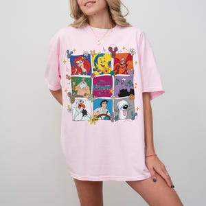 May include: A light pink t-shirt featuring a colorful graphic of characters from Disney's The Little Mermaid. The graphic includes Ariel, Flounder, Sebastian, King Triton, Ursula, and Prince Eric. The shirt is a crew neck style.