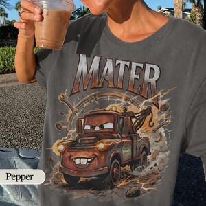 May include: A gray t-shirt featuring the word "MATER" in large, distressed lettering above a cartoon image of Mater the tow truck. The graphic includes lightning bolts and tools, creating a dynamic design.