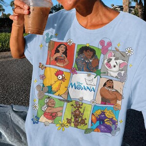 Op de afbeelding: Lichtblauw T-shirt met een kleurrijke afbeelding van personages uit de Disney-film Moana. Het ontwerp bevat afbeeldingen van Moana, Maui, Pua, Hei Hei en anderen, met het logo van de film in het midden.