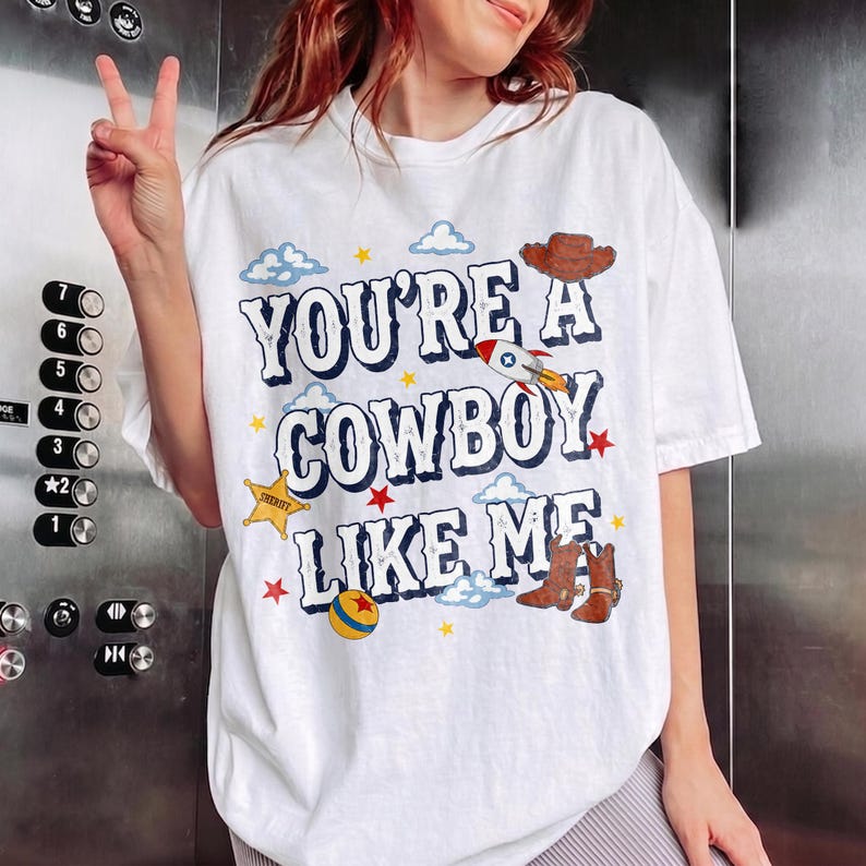 Puede incluir: Una camiseta blanca con el texto azul "YOU'RE A COWBOY LIKE ME" rodeado de gr&aacute;ficos coloridos que incluyen un sombrero de vaquero marr&oacute;n, un cohete, una estrella de sheriff amarilla, una pelota multicolor y botas de vaquero marrones con nubes y estrellas.
