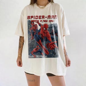May include: An off-white short-sleeve t-shirt featuring a large graphic print of Spider-Man in various action poses. The text "SPIDER-MAN" and "BRAND A NEW DAY" is printed in red above the character illustrations set against a dark city background.