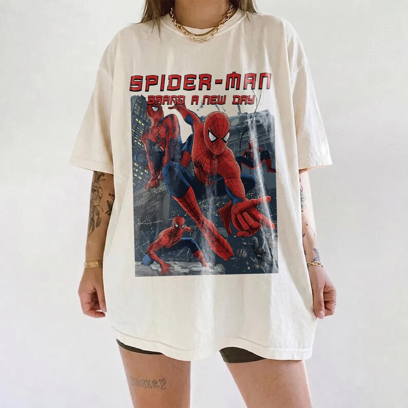 May include: An off-white short-sleeve t-shirt featuring a large graphic print of Spider-Man in various action poses. The text "SPIDER-MAN" and "BRAND A NEW DAY" is printed in red above the character illustrations set against a dark city background.