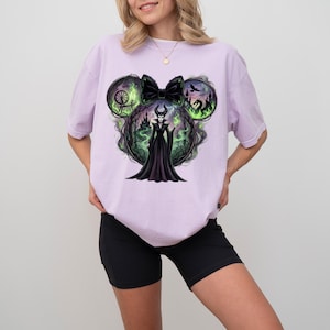 May include: A light purple t-shirt featuring a graphic of a Disney villain. The design includes a black bow, a silhouette of a castle, a dragon, and the villain in a black dress. The design is surrounded by green smoke.