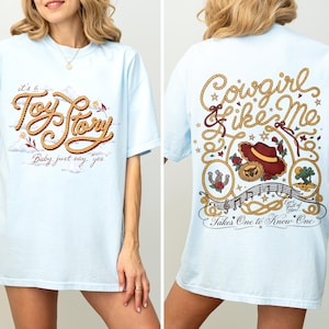 May include: A light blue t-shirt featuring a front graphic with rope-style text reading "it's a Toy Story Baby just say yes" and a back graphic with rope borders, a cowboy hat, and text reading "Cowgirl like Me Takes One to Know One".