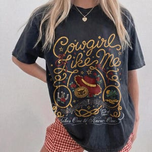 May include: A charcoal gray short-sleeve t-shirt featuring a graphic print with yellow rope lettering that reads Cowgirl Like Me and Takes One to Know One. The design includes a red cowboy hat, a horseshoe, roses, and musical notes.
