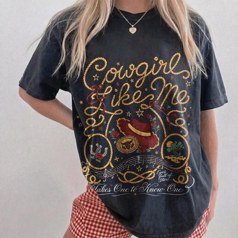 May include: A charcoal gray short-sleeve t-shirt featuring a graphic print with yellow rope lettering that reads Cowgirl Like Me and Takes One to Know One. The design includes a red cowboy hat, a horseshoe, roses, and musical notes.