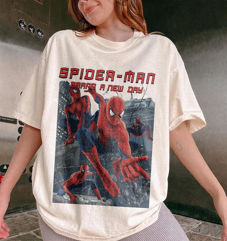 May include: A cream-colored short-sleeve t-shirt featuring a graphic print of Spider-Man in various action poses. The text "SPIDER-MAN" and "BRAND A NEW DAY" is printed in red above the character illustrations set against a dark city background.