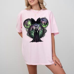 May include: Light pink t-shirt featuring a graphic of a Disney villain in a black dress, surrounded by green and black imagery. The design is shaped like Mickey Mouse ears with a black bow. The shirt is a relaxed fit.