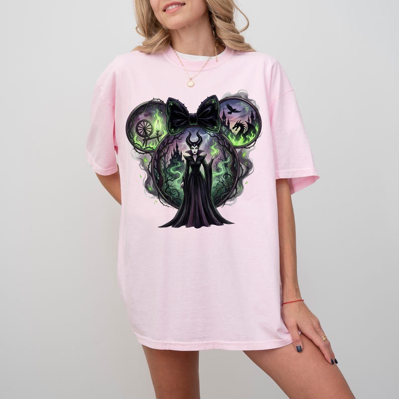 May include: Light pink t-shirt featuring a graphic of a Disney villain in a black dress, surrounded by green and black imagery. The design is shaped like Mickey Mouse ears with a black bow. The shirt is a relaxed fit.