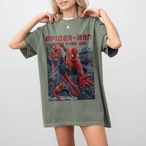 May include: An olive green short-sleeve t-shirt featuring a graphic print of the character Spider-Man in various action poses. The text "SPIDER-MAN" and "BRAND A NEW DAY" is printed in red above the character illustrations on the front of the shirt.