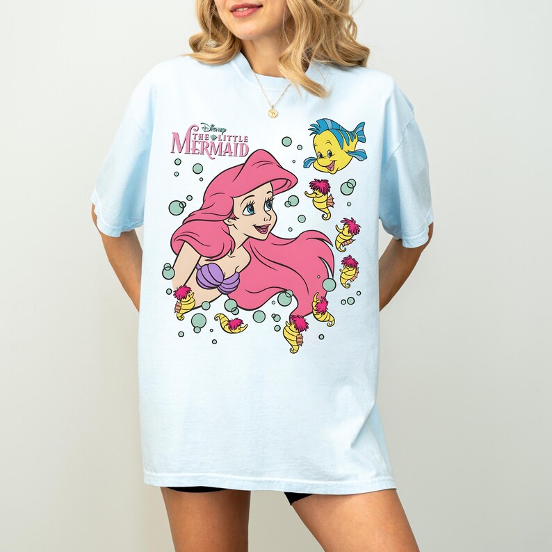 May include: Light blue t-shirt featuring a graphic of Ariel from Disney's The Little Mermaid. Ariel has pink hair and a purple seashell top. The shirt also includes Flounder and seahorse characters, with the text "Disney The Little Mermaid".