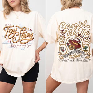 May include: An off-white short-sleeve t-shirt featuring a front graphic with rope-style text reading "it's a Toy Story" and "Baby just say yes". The back graphic displays "Cowgirl Like Me" with a cowboy hat, horseshoe, and musical notes.