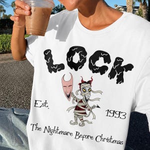 May include: White t-shirt featuring the word "LOCK" in a black, stylized font. Below the word is a cartoon illustration of a mummy-like character holding a mask. The text "Est. 1993 The Nightmare Before Christmas" is also printed on the shirt.