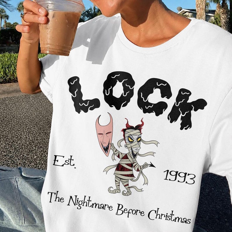 May include: White t-shirt featuring the word "LOCK" in a black, stylized font. Below the word is a cartoon illustration of a mummy-like character holding a mask. The text "Est. 1993 The Nightmare Before Christmas" is also printed on the shirt.