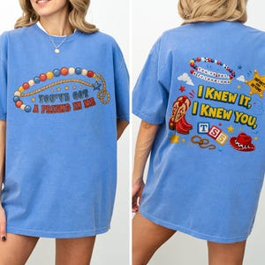 Pu&ograve; includere: Una t-shirt blu a maniche corte con una grafica frontale di una corda di perline e il testo "YOU'VE GOT A FRIEND IN ME." Il retro mostra un braccialetto di perline, una stella da sceriffo, stivali da cowboy, blocchi con "TS5," un cappello e il testo "I KNEW IT, I KNEW YOU."