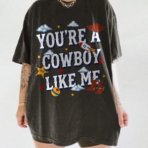 Puede incluir: Una camiseta gris carb&oacute;n de corte oversize con el texto blanco "YOU'RE A COWBOY LIKE ME" e ilustraciones gr&aacute;ficas coloridas de un sombrero de vaquero rojo, un cohete, una estrella de sheriff amarilla, botas marrones y nubes.