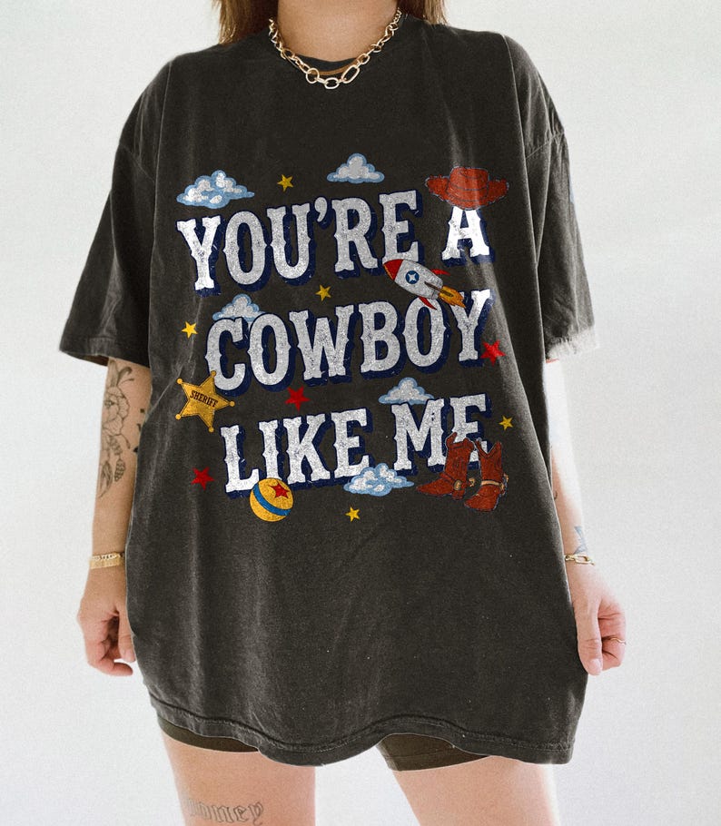 Puede incluir: Una camiseta gris carb&oacute;n de corte oversize con el texto blanco "YOU'RE A COWBOY LIKE ME" e ilustraciones gr&aacute;ficas coloridas de un sombrero de vaquero rojo, un cohete, una estrella de sheriff amarilla, botas marrones y nubes.