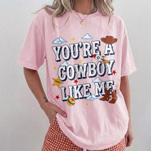 Puede incluir: Una camiseta de manga corta de color rosa claro con el texto YOU'RE A COWBOY LIKE ME en letras azul oscuro. El gr&aacute;fico incluye un sombrero de vaquero marr&oacute;n, un cohete, una estrella de sheriff amarilla, una pelota colorida, estrellas rojas y nubes azules.