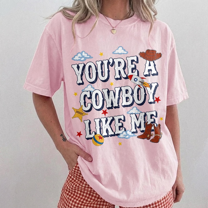Puede incluir: Una camiseta de manga corta de color rosa claro con el texto YOU'RE A COWBOY LIKE ME en letras azul oscuro. El gr&aacute;fico incluye un sombrero de vaquero marr&oacute;n, un cohete, una estrella de sheriff amarilla, una pelota colorida, estrellas rojas y nubes azules.