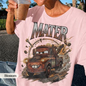 May include: A light pink t-shirt featuring a graphic of Mater from the Cars movie. The design includes the word "MATER" in a metallic font above the cartoon tow truck. The shirt has a crew neck and short sleeves.