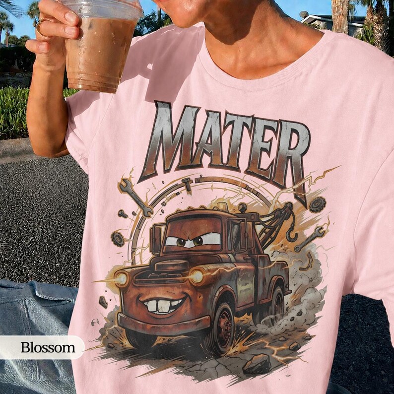 May include: A light pink t-shirt featuring a graphic of Mater from the Cars movie. The design includes the word "MATER" in a metallic font above the cartoon tow truck. The shirt has a crew neck and short sleeves.
