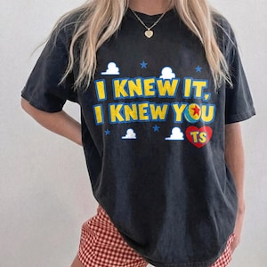 May include: A black short-sleeved t-shirt featuring the yellow text I KNEW IT, I KNEW YOU with a red heart containing the letters TS. The design includes small white clouds and blue stars, with the letter O replaced by a colorful ball graphic.
