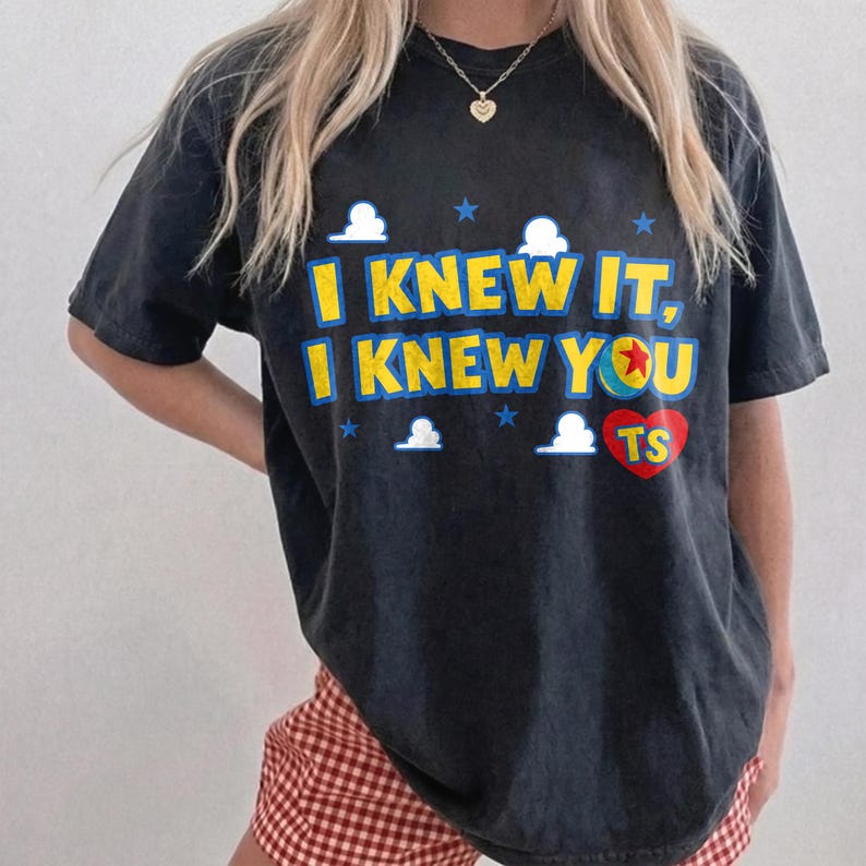 May include: A black short-sleeved t-shirt featuring the yellow text I KNEW IT, I KNEW YOU with a red heart containing the letters TS. The design includes small white clouds and blue stars, with the letter O replaced by a colorful ball graphic.