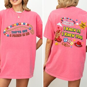 Pu&ograve; includere: Una t-shirt rosa acceso a maniche corte con una grafica frontale di una collana di perline colorate e il testo "YOU'VE GOT A FRIEND IN ME". Il retro mostra uno stivale da cowboy, una stella da sceriffo, un lazo e il testo "I KNEW IT, I KNEW YOU".