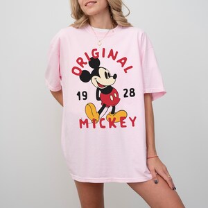 May include: A light pink t-shirt featuring a graphic of Mickey Mouse. The design includes the word "ORIGINAL" in red, the numbers "19" and "28", and the word "MICKEY" in red. Mickey Mouse is depicted in his classic outfit.
