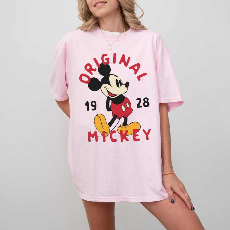 May include: A light pink t-shirt featuring a graphic of Mickey Mouse. The design includes the word "ORIGINAL" in red, the numbers "19" and "28", and the word "MICKEY" in red. Mickey Mouse is depicted in his classic outfit.