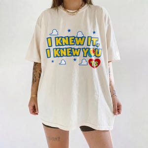 May include: An off-white short-sleeve t-shirt featuring the blue and yellow text I KNEW IT, I KNEW YOU with a red heart containing the letters TS. The design includes blue cloud and star graphics, with a gold chain necklace worn around the neck.
