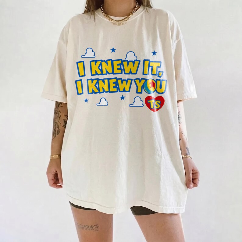 May include: An off-white short-sleeve t-shirt featuring the blue and yellow text I KNEW IT, I KNEW YOU with a red heart containing the letters TS. The design includes blue cloud and star graphics, with a gold chain necklace worn around the neck.