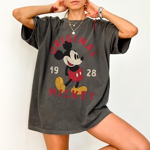 May include: Dark gray t-shirt featuring a distressed graphic of Mickey Mouse. The design includes the words "ORIGINAL MICKEY" and the numbers "1928" in red. The shirt has short sleeves and a relaxed fit.