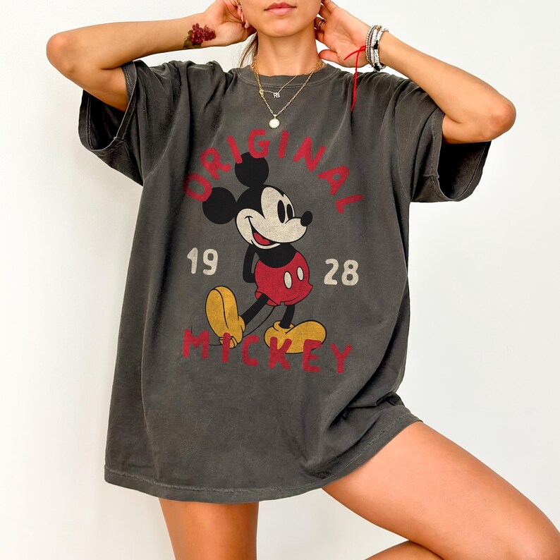 May include: Dark gray t-shirt featuring a distressed graphic of Mickey Mouse. The design includes the words "ORIGINAL MICKEY" and the numbers "1928" in red. The shirt has short sleeves and a relaxed fit.