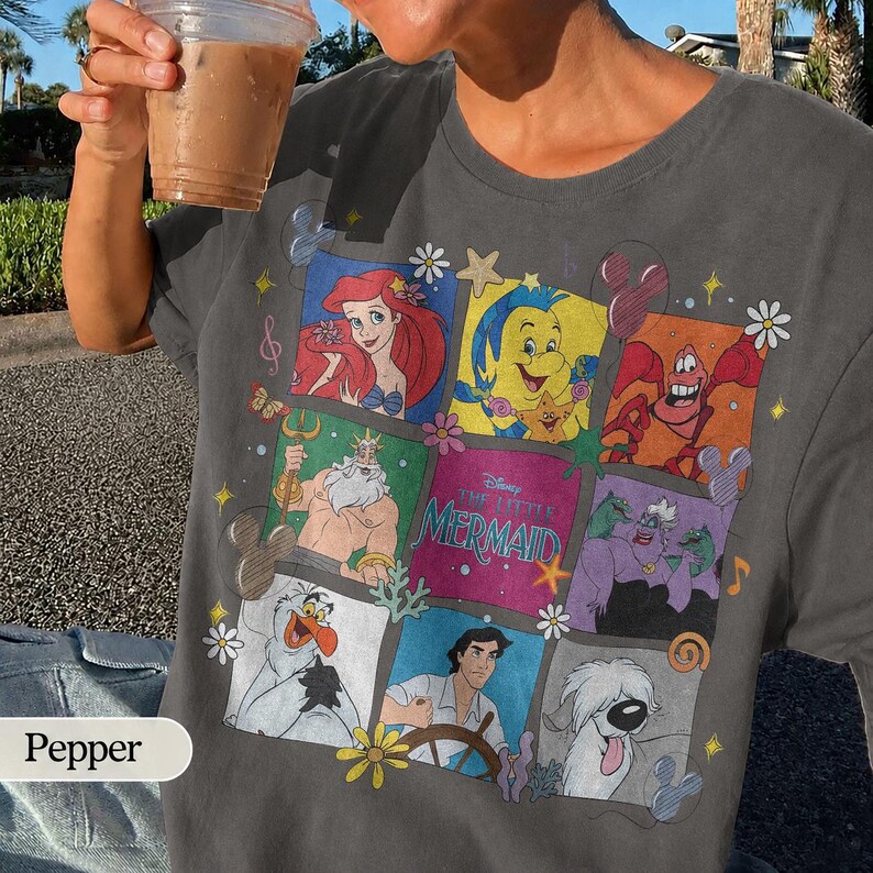 May include: Dark gray t-shirt featuring a colorful graphic of characters from Disney's "The Little Mermaid." The design includes Ariel, Sebastian, Flounder, and other characters in a grid pattern. The shirt has a crew neck and short sleeves.