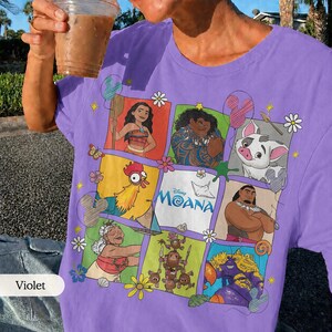 Op de afbeelding: Violetkleurig T-shirt met een kleurrijke collage van personages uit de Disney-film Moana. Het ontwerp bevat Moana, Maui, Hei Hei en Pua, met het logo van de film. Het shirt heeft een ronde hals en korte mouwen.