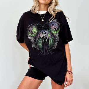 May include: Black t-shirt featuring a graphic of a Disney villain in a Mickey Mouse ear design. The design includes a bow, a castle, and a dragon. The graphic is in shades of green, black, and purple.