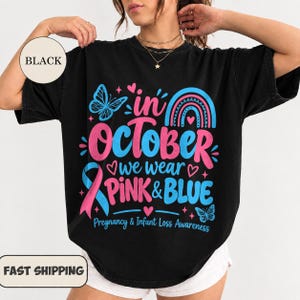 May include: A black t-shirt featuring a graphic design with the text In October We Wear Pink & Blue Pregnancy & Infant Loss Awareness, accompanied by a pink and blue awareness ribbon, a rainbow, butterflies, and small heart icons.