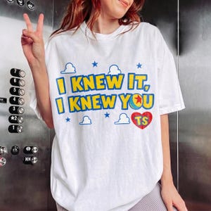 May include: A white short-sleeve t-shirt featuring the blue and yellow text I KNEW IT, I KNEW YOU, a colorful ball graphic, a red heart with the letters TS, and small blue cloud and star icons printed on the front of the garment.