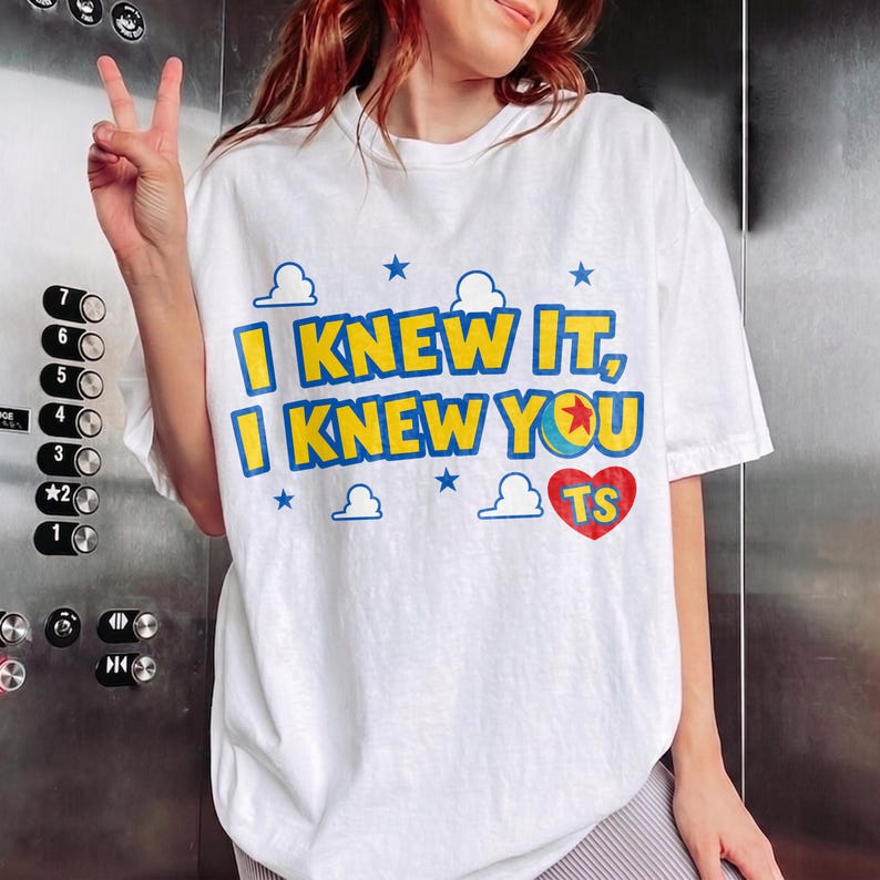 May include: A white short-sleeve t-shirt featuring the blue and yellow text I KNEW IT, I KNEW YOU, a colorful ball graphic, a red heart with the letters TS, and small blue cloud and star icons printed on the front of the garment.