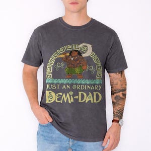 Puede incluir: Camiseta gris oscuro con un gr&aacute;fico de un personaje polinesio sosteniendo un arma, con el texto "JUST AN ORDINARY DEMI-DAD" en oro y verde. El dise&ntilde;o est&aacute; dentro de un arco decorativo.