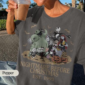 May include: Dark gray t-shirt featuring a graphic from "The Nightmare Before Christmas." The design includes Jack Skellington, Sally, and other characters, with the text "Nightmare Before Christmas EST. 1993."