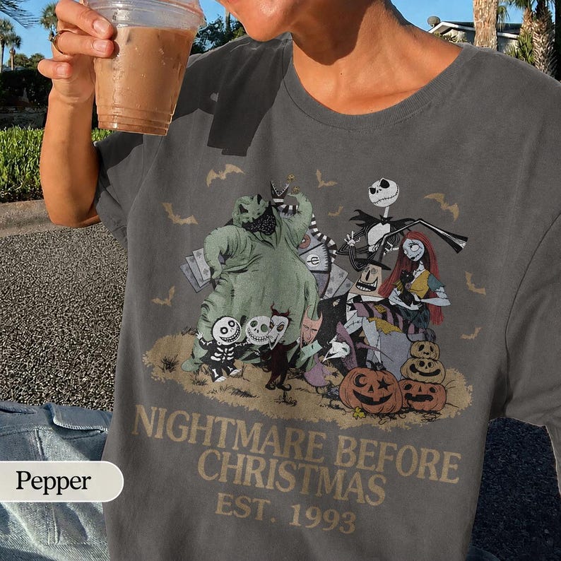 May include: Dark gray t-shirt featuring a graphic from "The Nightmare Before Christmas." The design includes Jack Skellington, Sally, and other characters, with the text "Nightmare Before Christmas EST. 1993."