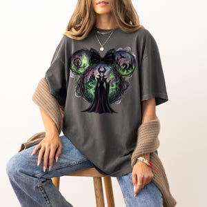 May include: Dark gray t-shirt featuring a graphic of a Disney villain in a Mickey Mouse ear design. The design includes a black bow, a castle, a dragon, and a spinning wheel, all in shades of green and black. The shirt is a loose fit.