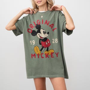May include: Olive green t-shirt featuring a vintage Mickey Mouse graphic. The design includes the word "ORIGINAL" in red, the numbers "19 28", and the name "MICKEY" in yellow. The shirt has short sleeves and a relaxed fit.